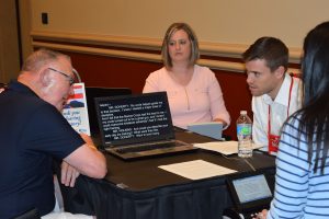 Four people sit around a table in a hotel conference room. In the back is a middle-aged woman concentrating while transcribing on a steno machine. On the left and right are two men -- one younger, one older, in conversation. In the foreground is the back of a yong woman; her steno machine is in front of her and a laptop is on the table with the spoken words appearing on the screen in real time.