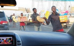 View is through the windshield of a car as if sitting in the passenger seat. A line of street vendors walk along the car holding various wares for sale. They are looking ahead.