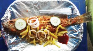 Fried whole fish covered with sliced cucumbers, sliced onions, and french fries with a side of ketchup. Next to the food is a set of plasticware. The plate is covered in aluminum foil.