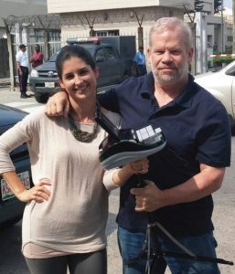 A young woman and an older man are facing the camera with their arms congenially around each other's shoulders as if friends. The woman is holding a steno machine on a tripod.