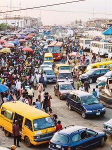 A street scene in Lagos: A narrow paved street with a line of cars (sometimes single file, sometimes double file), cars parked or waiting to move on either side of the street, pedestrians crowded mostly on the left side. Near the background, a cluster of colorful umbrellas. In the back, white nondescript buildings. At the top in the foreground and background are electrical wires.