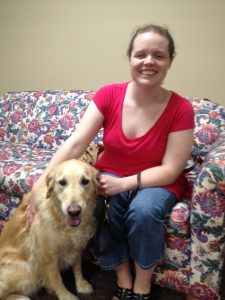 A smiling young adult woman, dressed cassually, sits on a floral couch with a golden retriever at her side.