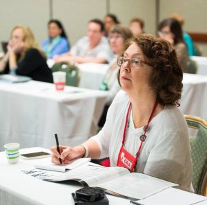 A middle-aged white woman listens attentively during a workshop while taking notes.