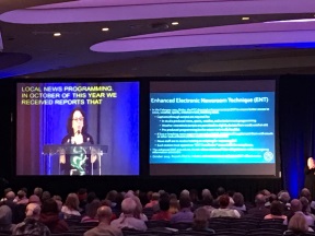 Rear view of a dark conference room filled with people looking toward two screens; one has the speaker with captions overlaid on top and the other has a slide from the presentation