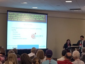 Rear view of a conference room with a man and woman presenting at the front. On the left is a screen with the PowerPoint presentation and captions overlaid on top.