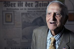 An elderly man is in the middle of speaking. He sits in front of a German newspaper projected behind him.