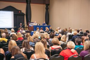 View from the back of a meeting room with rows of people facing a panel and a projector