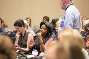 A woman speaks into a microphone. She is sitting amongst rows of people at a conference session.