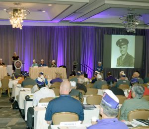 Back view of a conference room with a seated audience -- mostly men and some wearing commemorative military service hats. A woman stands at the podium in the front of the room. On the projector is a black and white photographer of a smiling young man in uniform, probably circa the 1940s