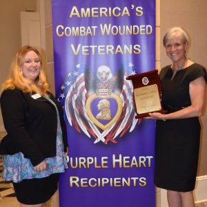 Two women, one holding a plaque in the shape of a scroll, stand in front of a banner reading "America's Combat Wounded Veterans -- Purple Heart Recipients." The wording is wrapped around an image of the Purple Heart medal in front of a bald eagle whose wings turn into the American flag.