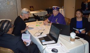 Four people sit around a table -- two are in coversation while the other two write the conversation on a steno machine and provide captioning