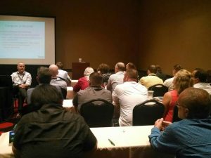 Back view of a packed classroom. In the front left, a man sits on a chair in front of a PowerPoint presentation; the slide is on the topic "computer as recorder."