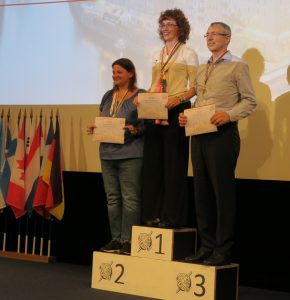 Three smiling people stand on a podium of various heights (1st, 2nd, and 3rd) holding certificates. In the background are a collection of international flags.