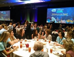 A large luncheon in a hotel ballroom with people seated at round tables; in the background is the logo for the 2017 NCRA Convention & Expo