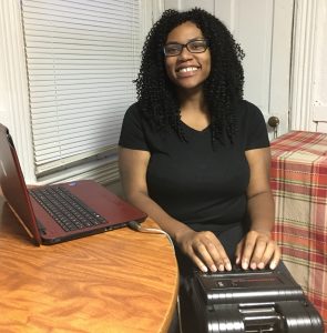 A young woman sits in front of a steno machine poised as ready to write. Her laptop is open on the table in front of her.