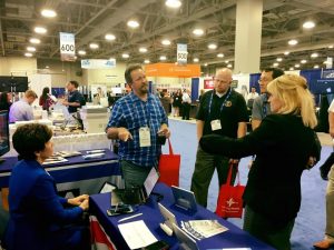 Set in a moderately busy vendor hall, two women in professional garb speak with a few men who are visiting the booth. One of the women is seated at a steno machine. On the table are flyers and propped up iPads.