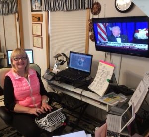 A woman sits in front of a steno machine, set up to work from home. On her desk is her laptop and paper notes propped up for easy viewing. On the wall is a television screen with a news show.