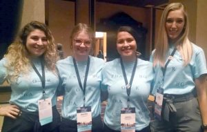 Four young women pose in matching light blue shirts with steno written on the front