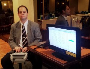 Man in a suit sitting at a steno machine next to a screen