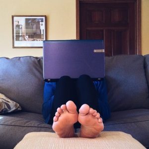 Front view of a person sitting barefoot on a couch with their laptop on their knees, blocking their face