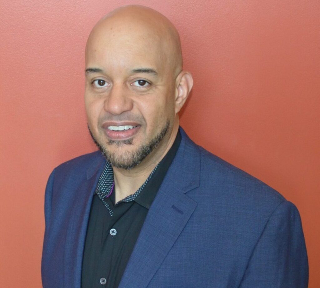 A man wearing a dark blue blazer over a black collared shirt smiles at the camera while standing in front of a solid orange-red background. He is bald with a neatly trimmed beard and goatee, presenting a polished and professional appearance.