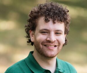 Smiling person with short curly hair, a trimmed beard, and a nose ring, wearing a green collared shirt, standing outdoors with a softly blurred natural background.