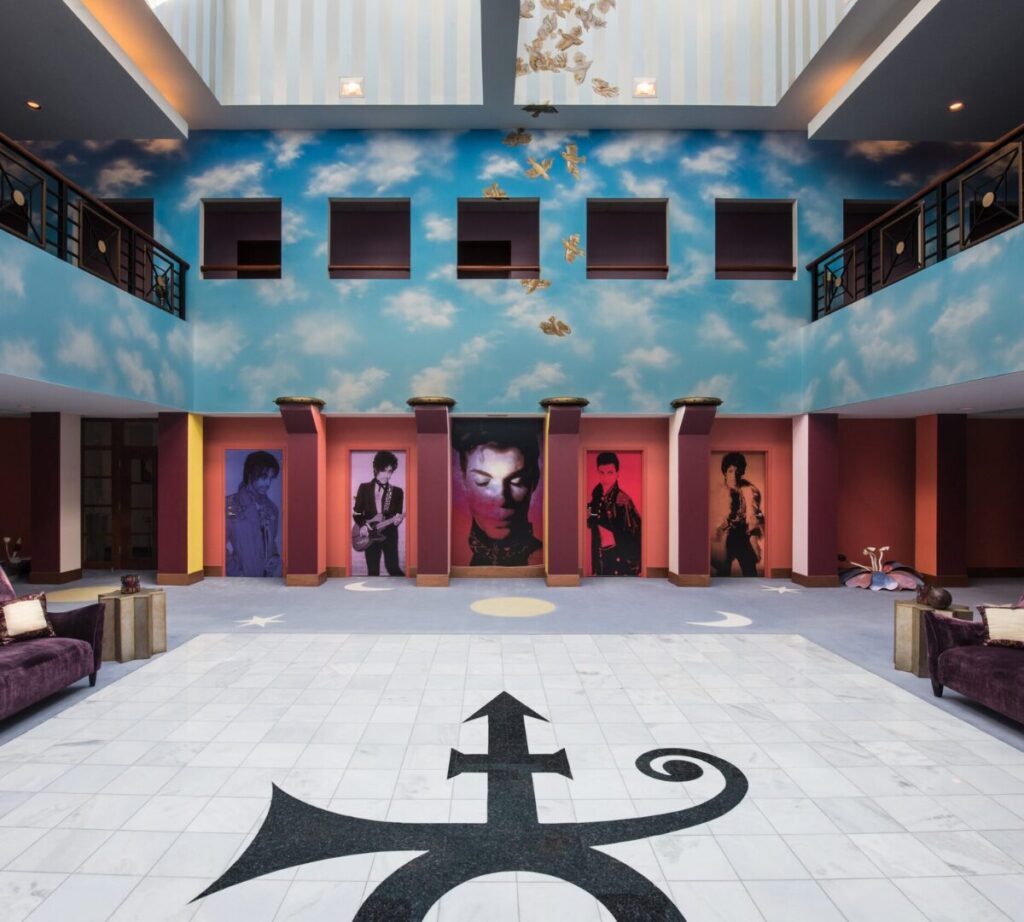 Alt text: A vibrant atrium inside Paisley Park, Prince’s former home and studio, featuring a sky-blue mural with clouds on the upper walls, gold doves suspended from the ceiling, and five large portraits of Prince in various iconic looks. The floor has a large black symbol of Prince's "Love Symbol" on white tiles. The room is flanked by plush purple seating and accented with celestial motifs like moons and stars.