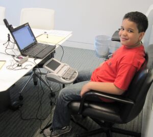 A young boy sits in a black office chair, smiling at the camera while using a steno machine connected to a laptop on a nearby table. The setup includes various cords, earphones, and writing materials, suggesting he is practicing or learning stenography in a professional or educational setting.