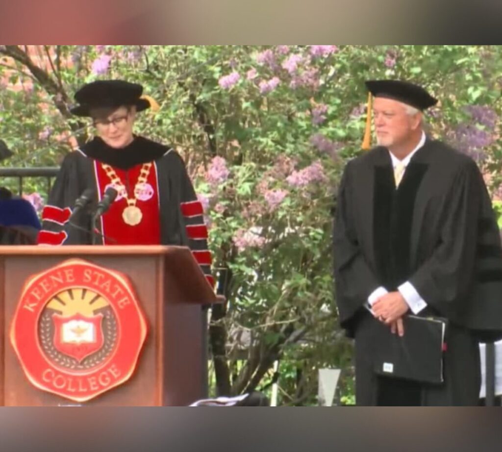 A woman in academic regalia speaks at a podium adorned with the Keene State College seal during an outdoor commencement ceremony. A man in similar regalia stands beside her, holding a folder. Both are under a canopy of blooming lilac bushes.