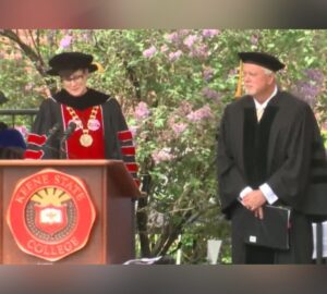 A woman in academic regalia speaks at a podium adorned with the Keene State College seal during an outdoor commencement ceremony. A man in similar regalia stands beside her, holding a folder. Both are under a canopy of blooming lilac bushes.