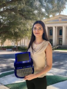 Woman standing in front of a school building holding a steno machine and smiling at the camera