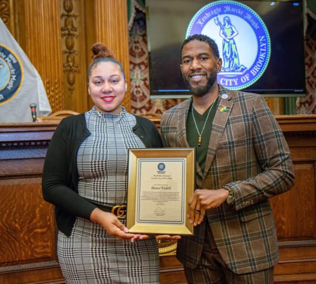 "Denee Vadell, wearing a black and white checkered dress and black sweater, stands next to a man in a brown plaid suit. They are smiling and holding a framed award certificate. The photo is taken inside a formal chamber with ornate wood paneling, flags, and a screen in the background displaying the City of Brooklyn seal.