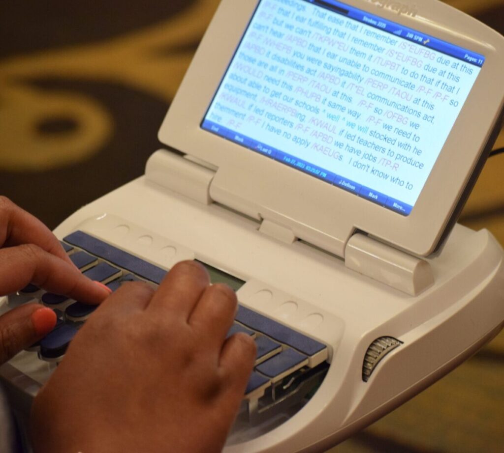A person types on a stenograph machine with a display screen showing real-time transcription in English text.