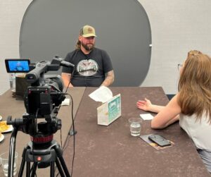 A man wearing a black T-shirt and tan baseball cap sits for an interview across from a woman with long reddish-brown hair. A professional video camera is set up on a tripod recording the conversation. On the table are glasses of water, a tissue box, a phone, and printed materials. The setting appears to be a quiet interview space with a gray backdrop behind the man.