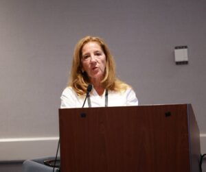 A woman with shoulder-length light brown hair speaks at a podium during a conference session. She wears a white blouse and a black lanyard, and a microphone is positioned in front of her.