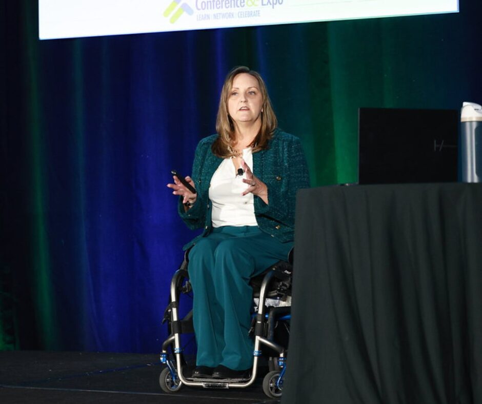A woman in a teal jacket and slacks speaks on stage from a wheelchair at a conference. She gestures with her hands while presenting beside a podium and laptop, with a dark blue and green curtain backdrop.