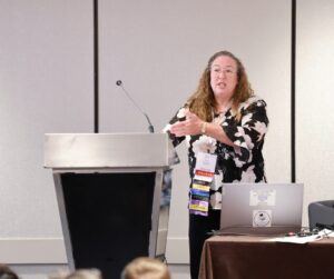 A woman wearing a floral blouse speaks at a podium during a conference session. She gestures with her hands while addressing the audience. Her name badge identifies her as a speaker with multiple NCRA credentials, including RDR and CRC. A laptop with a “Proud Stenographic Reporter” sticker sits on a nearby table.