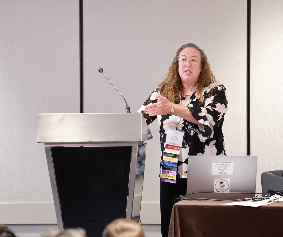 A woman wearing a floral blouse speaks at a podium during a conference session. She gestures with her hands while addressing the audience. Her name badge identifies her as a speaker with multiple NCRA credentials, including RDR and CRC. A laptop with a “Proud Stenographic Reporter” sticker sits on a nearby table.