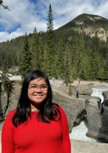 Woman in a red sweater in glasses standing in front of a river, smiling at the camera