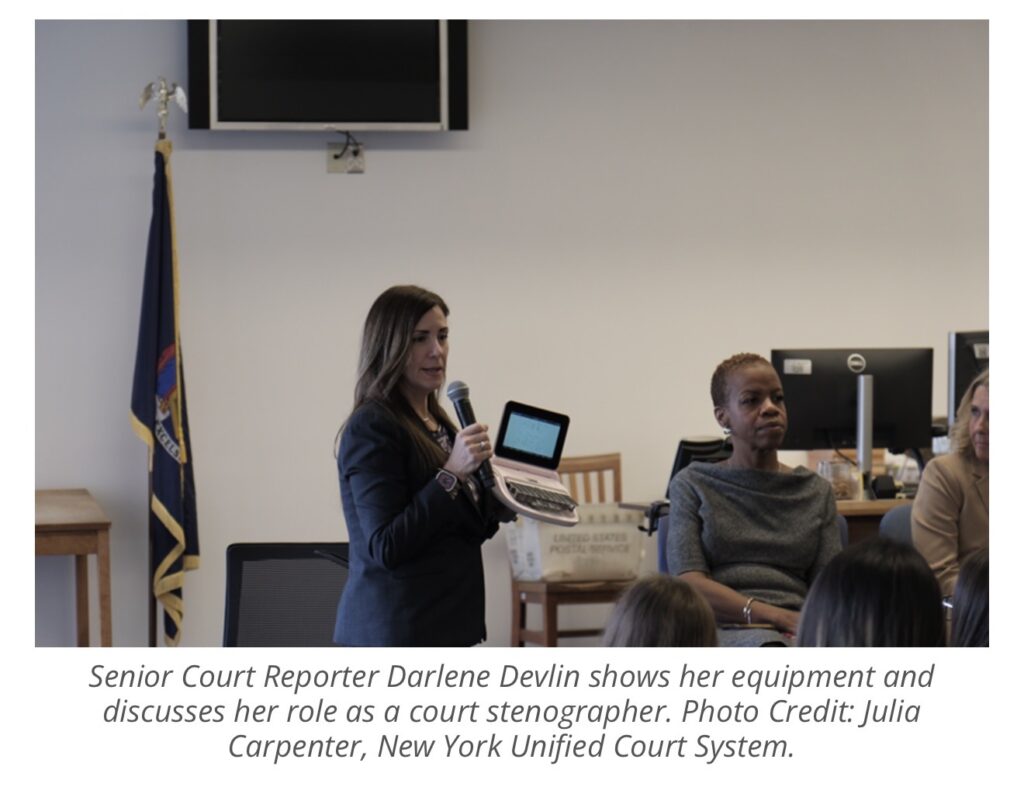 A woman standing holding a microphone and a steno machine presenting to a crowd