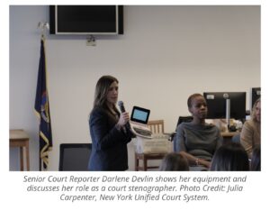A woman standing holding a microphone and a steno machine presenting to a crowd