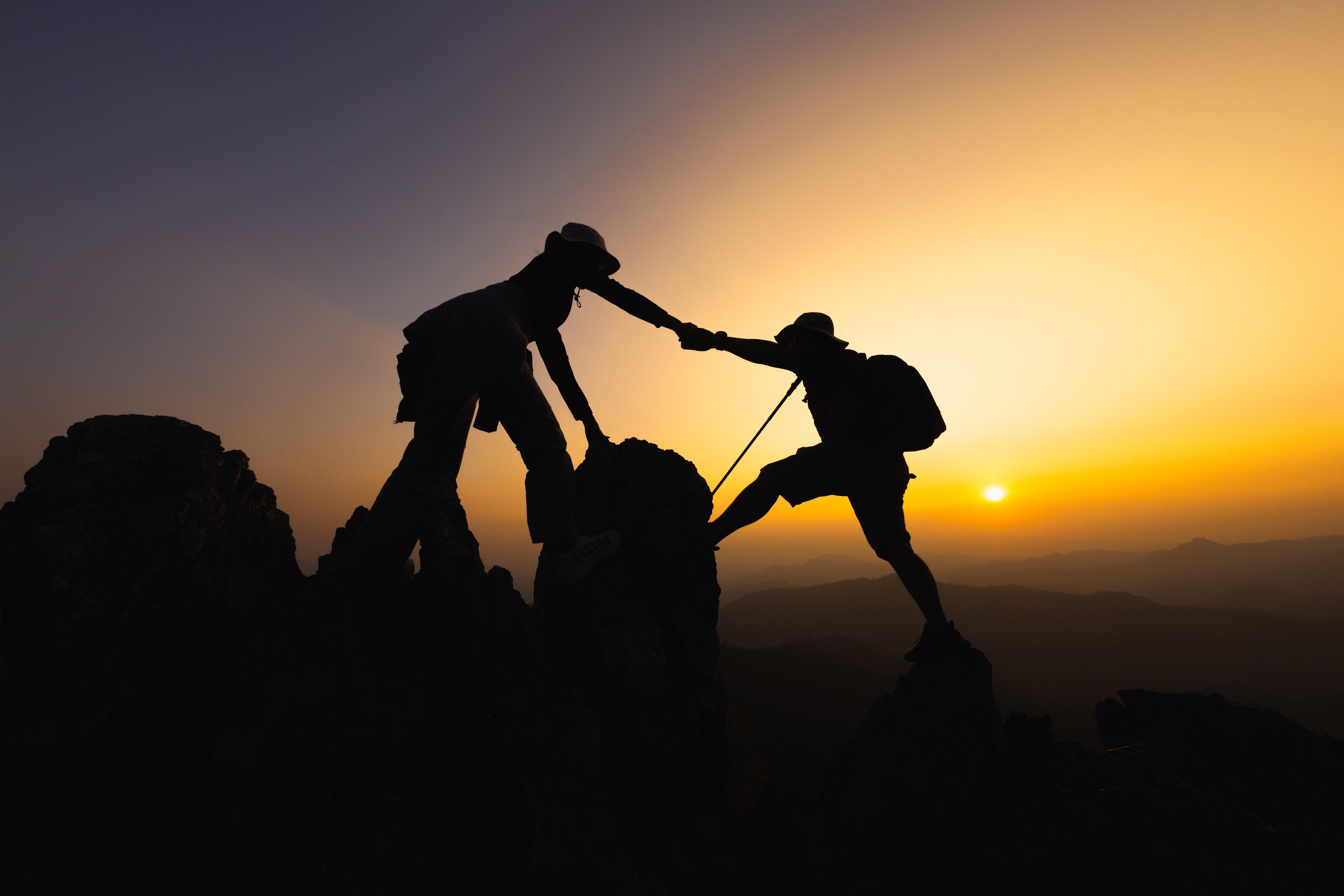 a silhouette of two people helping each other on a hike at sunset